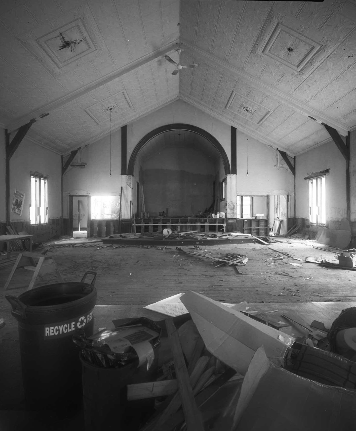 Interior of an abandoned meeting hall, ceiling tiles decaying above a debris-strewn floor.